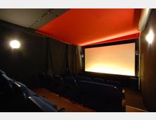 Kleiner Kinosaal mit dunklen Wänden, roten Deckenpaneelen und einer beleuchteten Leinwand im Hintergrund. Die Sitzreihen bestehen aus blauen Polstersesseln, und die Beleuchtung ist gedämpft.