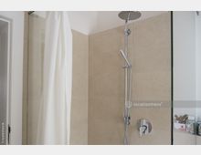 Dusche mit beigefarbenen Fliesen, einer Regendusche und Handbrause aus Edelstahl, einem weißen Duschvorhang und einer teilweisen Glasabtrennung. Im Hintergrund ist eine kleine Ablage mit Pflegeprodukten sichtbar.