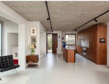 Modern open-plan kitchen and living area with a raw concrete ceiling, white walls, and black track lighting. Warm wood cabinetry and a peninsula with metal stools face a window and open door to greenery, with minimal furnishings and framed artwork along the wall.