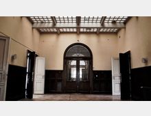 Ein leerer Raum mit hohen Decken und einer Glasziegeldecke, die Licht einlässt. Die Wände sind beige, der untere Wandbereich ist mit dunklem Holz getäfelt, und in der Mitte befindet sich eine doppelflügelige Tür mit einem halbkreisförmigen Oberlicht aus Glas.