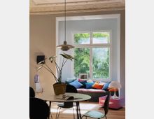 Wohnzimmer mit moderner Einrichtung, bestehend aus einem runden Tisch mit Stühlen, einem großen Fenster mit Blick auf grüne Bäume und einer gemütlichen Sitzbank mit bunten Kissen. Im Vordergrund ist eine Zimmerpflanze auf dem Tisch, daneben eine offene Zeitschrift, und im Hintergrund befinden sich ein kleiner rosa Beistelltisch sowie eine dekorative Lampe.