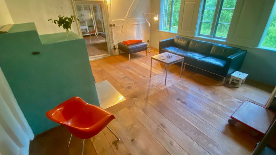 Gemütliches Wohnzimmer mit breiten Holzdielen, weiß verputzten Wänden und großen, grün gestrichenen Sprossenfenstern mit Blick ins Grüne. Ein schwarzes Ledersofa mit kleinem Couchtisch, eine Ottomane sowie ein gemauerter grüner Ofen und ein Durchgang in einen Nebenraum prägen den Raum.
