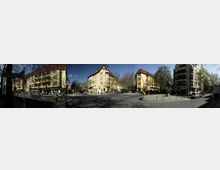 Straßenszene in einer Stadt mit gelben Wohngebäuden im Altbaustil und roten Dächern, umgeben von Bäumen. Im Vordergrund eine Kreuzung mit geparkten Autos, Fahrrädern, Fußwegen und einem "U-Bahn"-Schild auf der linken Seite.