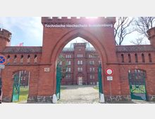Backstein-Eingangsportal mit Spitzbogen und der Aufschrift „Technische Hochschule Brandenburg“, dahinter ein historisches, mehrstöckiges Gebäude mit symmetrischen Fenstern und Türmen. Das Gelände ist von Grünflächen und Kopfsteinpflaster umgeben.