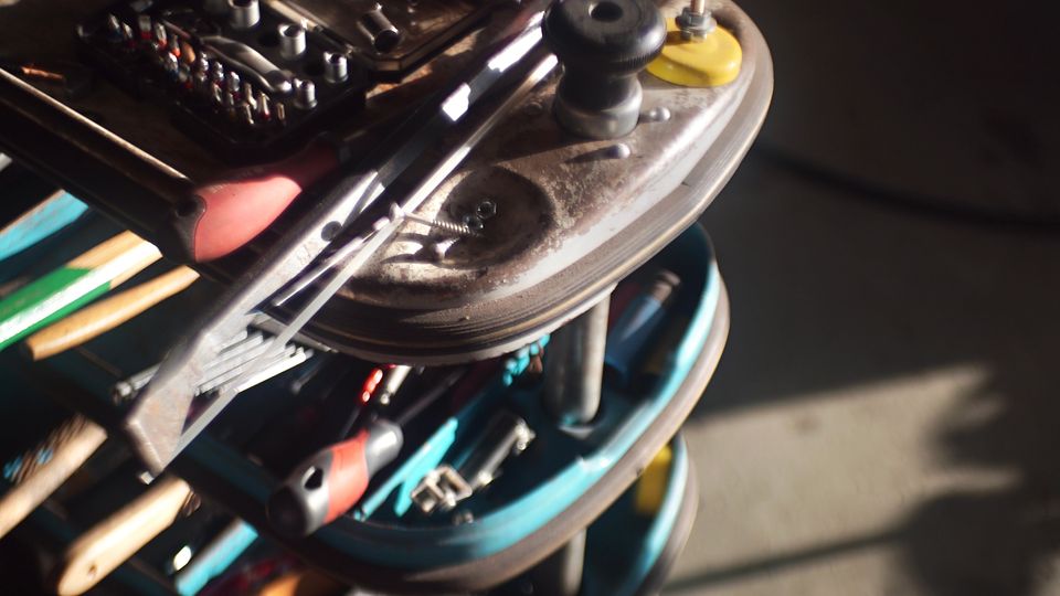 Werkstatt- oder Garagenbereich mit einem rollbaren Werkzeugwagen, auf dem Handwerkzeuge, Bitsätze, Schraubendreher und eine Spraydose liegen. Im Vordergrund ist eine Metallrolle mit Seil bzw. Kabel zu sehen, darunter mehrere Ablagefächer mit weiterem Werkzeug auf Betonboden.