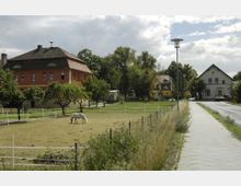 Ländliche Straßenszene mit einem eingezäunten Feld im Vordergrund, auf dem ein weißes Pferd grast. Im Hintergrund stehen ein rotes Wohngebäude, weitere Häuser, Bäume und ein Gehweg entlang einer schmalen, asphaltierten Straße.