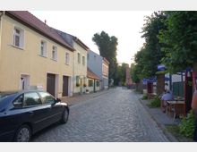 Pflastersteinstraße mit aneinandergereihten zweistöckigen Wohnhäusern in hellen Farben auf der linken Seite und schattigen Bäumen sowie Café-Tischen auf dem Bürgersteig rechts. Im Hintergrund ist eine Baumreihe und ein Weg sichtbar, der in die Ferne führt.