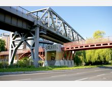 Eine stählerne Eisenbahnbrücke in Berlin, ergänzt durch eine rote Fußgängerbrücke und eine Betonstütze mit dem Schriftzug „Berlin“. Im Hintergrund sind Bäume, ein Straßenabschnitt und Teile des Deutschen Technikmuseums zu sehen.