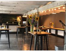 Veranstaltungsraum mit Holztischen, die mit bunten Blumenvasen dekoriert sind. Im Hintergrund eine Theke mit Gitterelementen und Spiegeln, die von warmem Licht beleuchtet wird, sowie eine graue Wand.