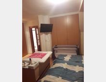 Ein Schlafzimmer mit einem Bett im Vordergrund, einem Wickeltisch links, einem zusammenklappbaren Kinderbett rechts und einem Flachbildschirm, der an der Wand befestigt ist. Im Hintergrund befinden sich ein Kleiderschrank mit Holzfronten und eine Tür, die zu einem anderen Raum führt.