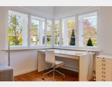 Heller Arbeitsbereich in einem Zimmer mit breiter Fensterfront über Eck und weißen Sprossenfenstern mit Blick ins Grüne. Unter den Fenstern steht ein schlichter weißer Schreibtisch mit Bürostuhl und Lampe, daneben ein schmaler Rollcontainer auf Holzdielenboden.