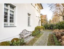 Seitenansicht eines weißen Wohnhauses mit Sprossenfenstern und hellem Putz. Im Vordergrund steht eine Holzbank auf einem kleinen gepflasterten Bereich neben einem Kiesweg, umgeben von Blumenbeeten und Sträuchern.