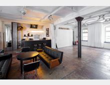 Geräumiger, moderner Innenraum mit einer Sitzecke aus schwarzen Ledermöbeln, einem kleinen runden Tisch und einer Bar mit Hockern im Hintergrund. Der Raum hat weiße Wände, Deckenbalken, große Fenster mit Vorhängen und einen Boden in rustikalem Beton-Look.