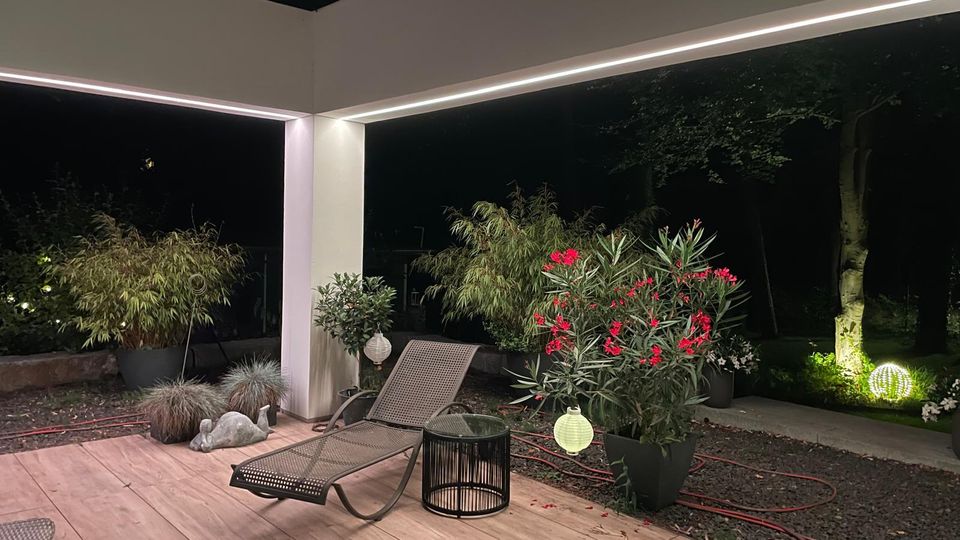 Überdachte Terrasse bei Nacht mit Holzdeck, weißer Stütze und integrierten LED-Lichtleisten entlang der Decke. Im Vordergrund stehen eine Liege und ein kleiner Beistelltisch, umgeben von großen Pflanzkübeln mit Bambus und blühenden Sträuchern sowie Gartenbeleuchtung im Hintergrund.