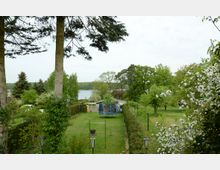 Ein grüner Garten mit Obstbäumen, blühenden Sträuchern und Hecken, im Hintergrund ein See mit umliegendem Wald. Im Vordergrund stehen zwei Bäume, eine blaue Trampolinstruktur und eine Wäschespinne.