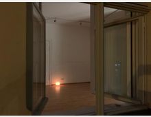 Blick durch ein offenes Fenster in einen leeren Raum mit Holzfußboden; eine kleine runde Lampe auf dem Boden beleuchtet den Raum schwach.