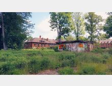 Eine verlassene Backsteinanlage mit langen Gebäuden und teilweise eingefallenen Dächern, umgeben von dichten Bäumen und einer verwilderten Wiese im Vordergrund. Rechts steht ein kleines, graffiti-bedecktes Betongebäude.