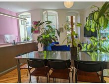 Gemütliche Wohnküche mit Holzboden, einem Esstisch mit Metalloberfläche und Stühlen im Vordergrund. Im Hintergrund befinden sich eine Küchenzeile mit rosa Wand, große Zimmerpflanzen, helle Fenster mit viel Tageslicht und ein Wohnzimmerbereich mit blauer Couch.