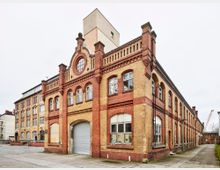 Backsteinbau eines ehemaligen Industriegebäudes mit roten und gelben Ziegelsteinen, Rundbogenfenstern und verziertem Giebel. Das Gebäude steht auf einem offenen Hof mit betonierter Fläche, daneben weitere ältere Backsteinstrukturen.