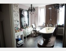 Innenansicht eines individuellen Badezimmers mit freistehender Badewanne auf einer gefliesten Plattform, zwei großen Fenstern, einem schmiedeeisernen Kronleuchter und Möblierung im Vintage-Stil. Neben einem Waschtisch sind zahlreiche Parfümflakons zu sehen, und ein großes Porträt hängt an der Wand.