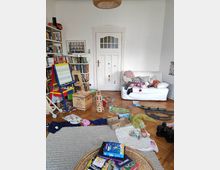 A cluttered living room with a white wooden door, a couch with scattered cushions, and shelving filled with books. The wooden floor is covered with toys, books, and game pieces, suggesting an active play area.