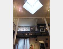 Innenansicht eines modernen Galerie- oder Museumsraums mit hoher Decke, einem großen Dachfenster und einer umlaufenden Empore mit Metallgeländern. Unten sind Vorhänge und beleuchtete Exponate sichtbar, während an der Empore Poster hängen.