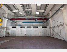 Industriehalle mit hohen Wänden und einer Tragkonstruktion aus Stahlträgern. Im Vordergrund ist ein leerer Boden mit roten Fliesen zu sehen, an der Stirnseite befinden sich mehrere Metalltüren und darüber eine rote Laufkatze mit einem Schienensystem und einer Tragkraftangabe von 5 Tonnen.