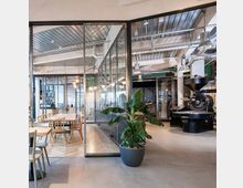 Ein modernes Café oder Workspace mit großen Glaswänden, einem langen Holztisch mit Stühlen im Vordergrund und einer Kaffeeröstmaschine im Hintergrund. Die Decke besteht aus sichtbaren Metall- und Lüftungselementen, und eine große grüne Zimmerpflanze steht zentral.