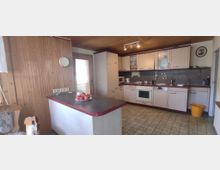 Küche mit Holzvertäfelung und Fliesenboden, ausgestattet mit hellen Einbauschränken, elektrischen Geräten wie einem Herd, einer Mikrowelle und einem Geschirrspüler. Im Vordergrund eine dunkle Arbeitsinsel mit einer Schale Äpfel, im Hintergrund eine Tür mit Milchglas.