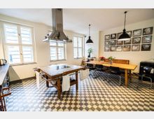 Helle Wohnküche mit großen Sprossenfenstern und schwarz-weiß kariertem Fliesenboden, in der Mitte eine Kochinsel aus Holz mit integrierter Herdplatte und markanter Dunstabzugshaube. Rechts steht ein langer Holztisch mit Stühlen und gepolsterter Bank unter schwarzen Pendelleuchten, an der Wand eine Reihe gerahmter Bilder.