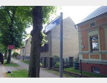 Gehweg einer Wohnstraße mit Bäumen, direkt angrenzenden Häusern und Vorgärten, darunter ein historisches Backsteinhaus mit grünen Fassadenelementen. Im Vordergrund ein Straßenschild und ein Verkehrsschild mit der Aufschrift „Achtung Feuerwehrausfahrt“.