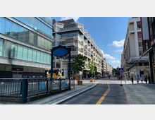 Straßenszene in einer Stadt mit modernen Gebäuden aus Glas und Stein auf beiden Seiten. Im Vordergrund eine mit Geländern eingefasste U-Bahn-Zugangsstruktur, dahinter eine Fahrbahn mit gelben Markierungen, Fußgänger, Bäume und Laternen.