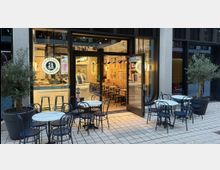 Außenansicht eines modernen Cafés mit halb geöffneten Glastüren, die den Innenraum mit Sitzbereichen und einer Theke zeigen. Draußen stehen rund um kleine weiße Tische schwarze Metallstühle auf einem gepflasterten Boden, eingerahmt von Olivenbäumen in großen Pflanzkübeln.