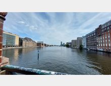 Blick auf einen breiten Fluss (Spree) in einer urbanen Umgebung, flankiert von modernen und historischen Gebäuden mit Backsteinfassaden. Im Vordergrund ein grünes Geländer mit Graffiti, im Hintergrund sind weitere Gebäude und ein hoher Turm zu sehen.