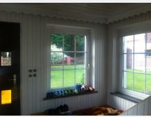 Ein Eingangsbereich mit weißer Holzvertäfelung, zwei großen Fenstern mit Blick in einen Garten und einer Haustür links. Auf den Fensterbänken und einer Bank im Raum stehen Schuhe und andere Gegenstände.