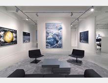 Moderner Galerieraum mit weißen Wänden, mehreren großformatigen Kunstwerken an den Wänden und minimalistischen Möbeln, darunter schwarze Stühle und ein grauer Couchtisch auf einem runden Teppich. Im Hintergrund ist ein Bürobereich sichtbar.