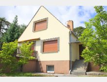 Einfamilienhaus mit gelber Fassade und braunem Sockel, rechteckigen Fenstern mit Rollläden und einem steilen Satteldach. Vor dem Haus befindet sich ein kleiner Garten mit Bäumen und einer gepflasterten Treppe, die zum Eingang führt.