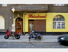 Straßenansicht einer alten Fleischerei mit gelben Fliesen und einem verschlossenen Rolltor. Vor dem Gebäude parken ein rotes Motorrad, ein blaues Moped und ein Auto; daneben steht eine grüne Straßenlaterne mit einem Plakat.