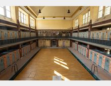 Historischer Innenraum eines ehemaligen Schwimmbads mit einem leeren Becken, das mit Holzplatten abgedeckt ist. Das Gebäude hat hohe Fenster, zweistöckige Umkleidekabinen mit Metalltüren und Backsteinwände, die den Raum strukturieren.