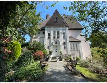 Großes, weißes Einfamilienhaus im traditionellen Stil mit steilem, braunem Satteldach und dekorativen Fenstern, umgeben von einem gepflegten Garten mit Wegen, Bänken und verschiedenen Bäumen und Pflanzen. Die Eingangstreppe führt zu einer verzierten Tür mit Laternen und Blumendekorationen.