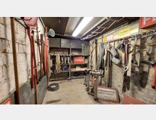 Lagerraum mit Feuerwehrausrüstung in einem historischen Stil: An den Wänden hängen Werkzeuge wie Äxte, Schläuche und Gurtbänder, zusätzlich sind Schilder mit Warnhinweisen angebracht. Im Raum befinden sich ein Holzfußboden, ein Regal mit Seilen und Objekten sowie eine alte Feuerlöschpumpe.