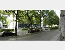 Straßenszene mit Bäumen, parkenden Autos und einem breiten Gehweg; im Hintergrund sind moderne Gebäude mit Glasfassaden zu sehen, die teilweise durch Grünflächen voneinander getrennt sind.
