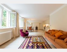 Helles Wohnzimmer mit offener Küche, ausgestattet mit einer orangefarbenen Couch, einem lila Sessel und einem Couchtisch auf einem farbenfrohen Teppich. Im Hintergrund befinden sich ein Esstisch mit Stühlen und eine moderne Einbauküche, während große Fenster auf der linken Seite Tageslicht hereinlassen.