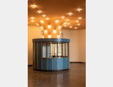 Runde Empfangskabine mit blauer Verkleidung und goldenen Akzenten, umgeben von einem offenen Raum mit glänzendem dunklem Boden; die auffällige Decke ist mit einer großen, säulenartigen Struktur und vielen integrierten Leuchten gestaltet.