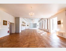 Heller Festsaal mit großzügigem Parkettboden, hohen Decken und einem Kristallleuchter. Die Wände sind weiß gestrichen und mit dekorativen Gemälden verziert; große Fenster mit Vorhängen auf der rechten Seite lassen viel Tageslicht herein.