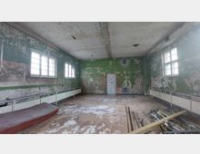 Ein leerer, heruntergekommener Raum mit grünen, stark abblätternden Wänden und mehreren rechteckigen Fenstern, die diffuses Licht einlassen. Der Boden ist verschmutzt, es liegen Baumaterialien herum, und an den Wänden befinden sich tiefe Putzschäden; im Hintergrund ist eine zugemauerte Tür zu sehen.