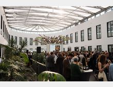 Geräumiger, moderner Veranstaltungsraum mit einer ovalen Glasdachkonstruktion, weißen Wänden mit rechteckigen Fenstern und einer zentralen Pflanzeninstallation in Baumform; am Rand des Raumes befinden sich Grünpflanzen und dazwischen sind Stehtische sichtbar.