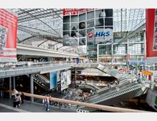 Moderne Bahnhofshalle mit mehreren Ebenen, Rolltreppen und Glasdach; im Hintergrund großflächige Werbebanner und Geschäfte. Menschen bewegen sich durch die hell beleuchtete Struktur mit sichtbaren Stahl- und Glaselementen.