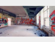 Verlassener Raum mit gefliesten Wänden, zahlreichen Graffiti und einem Boden, der mit Schutt bedeckt ist. Große Fenster an der rechten Wand lassen Tageslicht herein, während an der Decke unbenutzte Luftkanäle zu sehen sind.