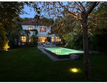 Beleuchteter Garten mit einem rechteckigen Pool in der Mitte, umgeben von Rasen und Bäumen; im Hintergrund steht ein zweistöckiges Wohnhaus mit weißen Wänden, roten Dachziegeln und großen Fenstern, die warm erleuchtet sind.